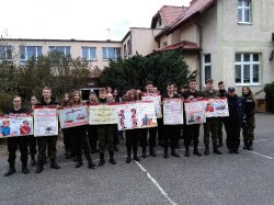 Fotografia kolorowa. Grupa kilkunastu uczniów klasy mundurowej stoi na boisku szkolnym i prezentuje plakaty dotyczące przeciwdziałania uzależnieniom