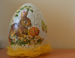 Fotografia kolorowa przedstawiająca ceramiczne jajo zdobione metodą dekupaż ustawione na żółtym sianku.