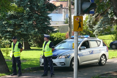 Zdjęcie w kolorze przedstawiające dwóch policjantów ruchu drogowego stojących wraz z radiowozem w rejonie przejścia dla pieszych.