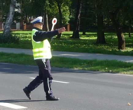 Zdjęcie kolorowe. Umundurowany policjant ruchu drogowego wydaje za pomocą tarczy sygnał do zatrzymania pojazdu