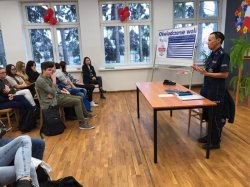 Fotografia kolorowa  obrazująca, jak profilaktyk społeczny chodzieskiej Policji prowadzi pogadankę z młodzieżą - ujęcie z boku.