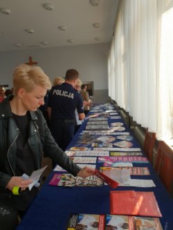 Zdjęcie w kolorze, na którym widać osoby, które uczestniczyły w  debacie, podczas oglądania materiałów profilaktycznych zgromadzonych na stole. W tle widoczny policjant, który rozmawia z obecnymi na debacie osobami.