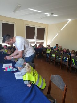 Fotografia kolorowa przedstawiająca policjanta, który pomaga dzieciom zrobić odbitki linii papilarnych. W tle widoczne przedszkolaki.